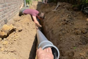 A person is lying in a trench next to a brick wall, working on connecting large gray pipes for underground plumbing—part of a sewer line repair. Another hand helps guide a pipe section into place. The scene is outdoors in daylight.