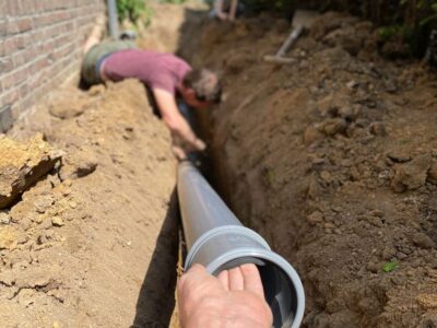A person is lying in a trench next to a brick wall, working on connecting large gray pipes for underground plumbing—part of a sewer line repair. Another hand helps guide a pipe section into place. The scene is outdoors in daylight.