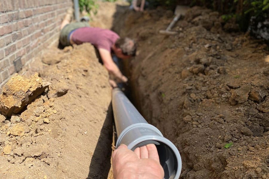 A person is lying in a trench next to a brick wall, working on connecting large gray pipes for underground plumbing—part of a sewer line repair. Another hand helps guide a pipe section into place. The scene is outdoors in daylight.